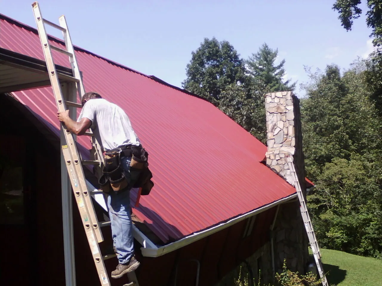 Roofer on a ladder beside a red metal roof and stone chimney — Soffit & Fascia Repair by Roofing Brothers in Forest